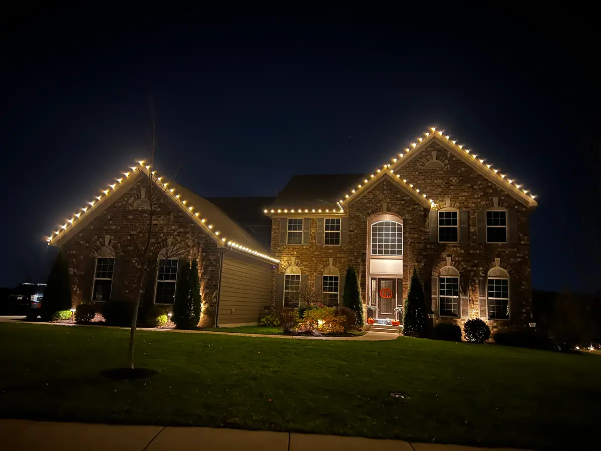 A stone-front two-story home traced in warm white lights with uplit landscaping